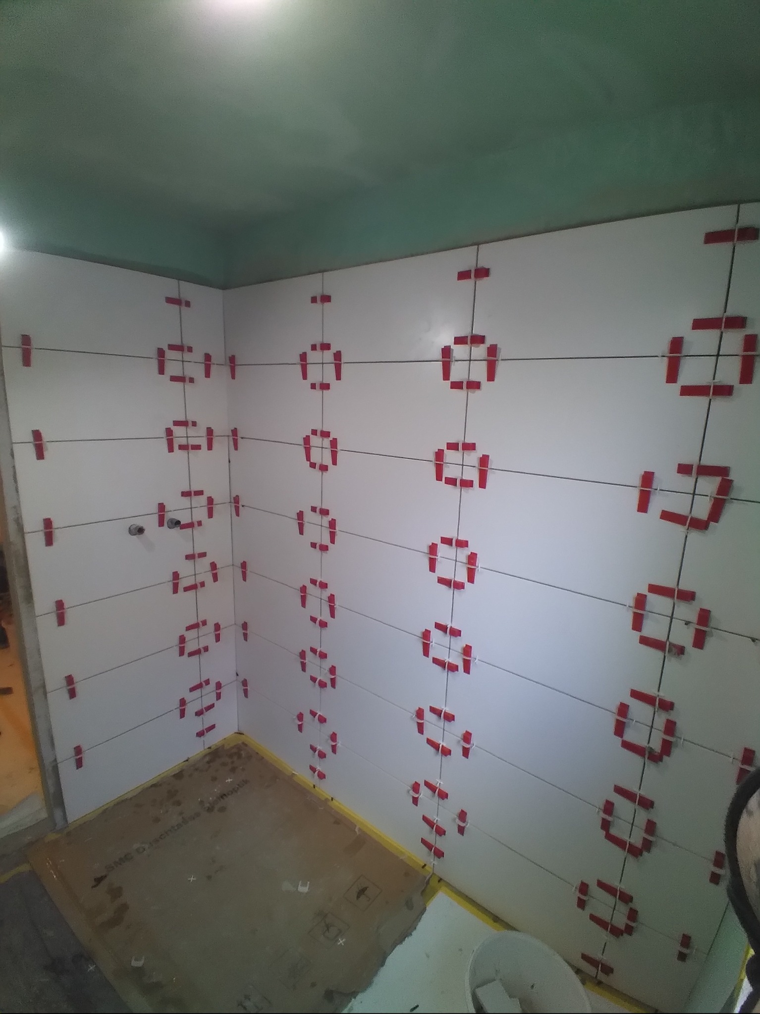 Białe płytki ścienne w łazience, ułożone z użyciem czerwonych klipsów i sznurka do poziomowania. Widoczne fragmenty podłogi i sufitu. Etap prac glazurniczych.
