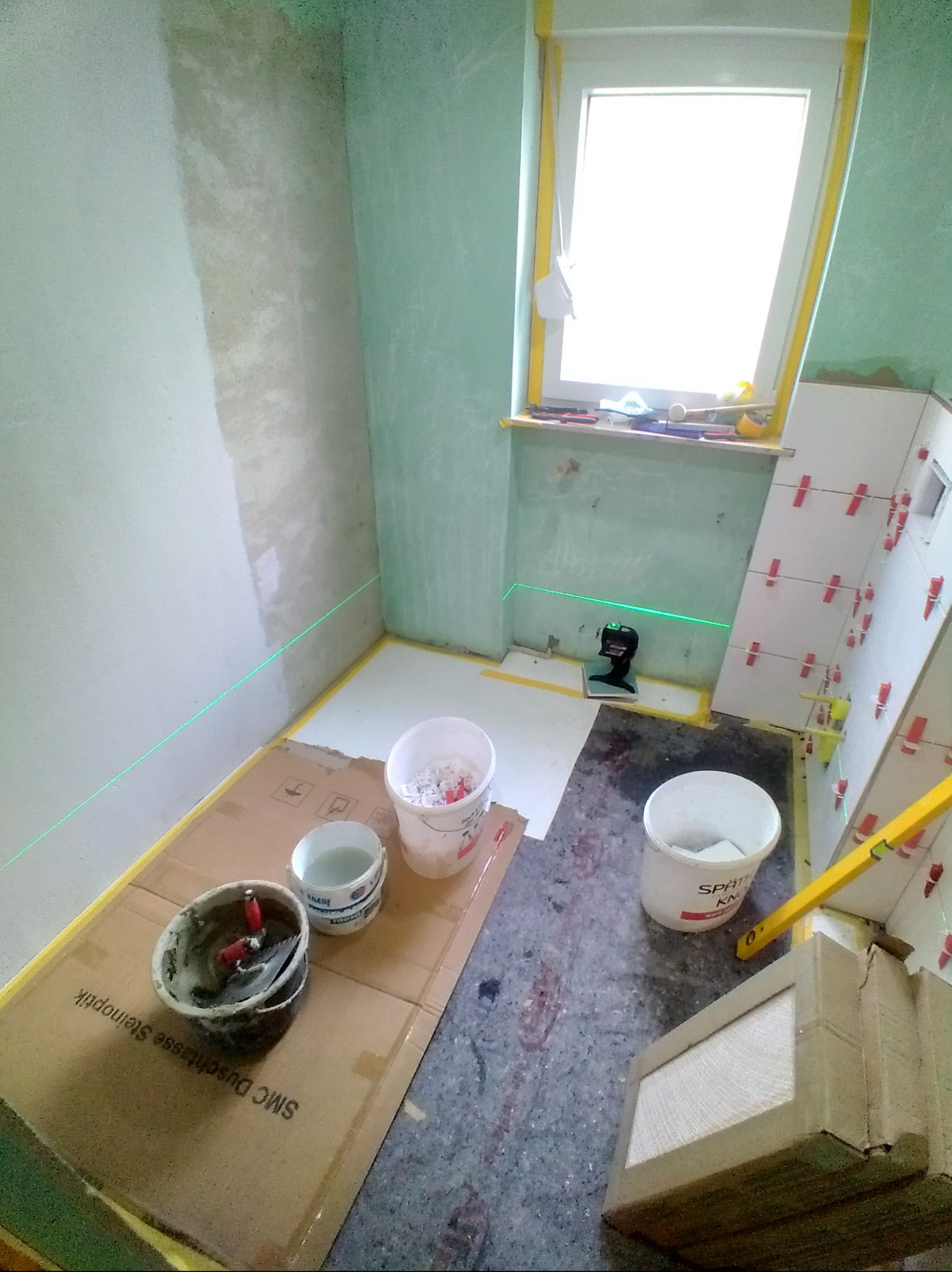 Remont łazienki w trakcie: laser krzyżowy, wiaderka z zaprawą, poziomica i płytki z krzyżykami dystansowymi. Przygotowanie do układania płytek w małej łazience.