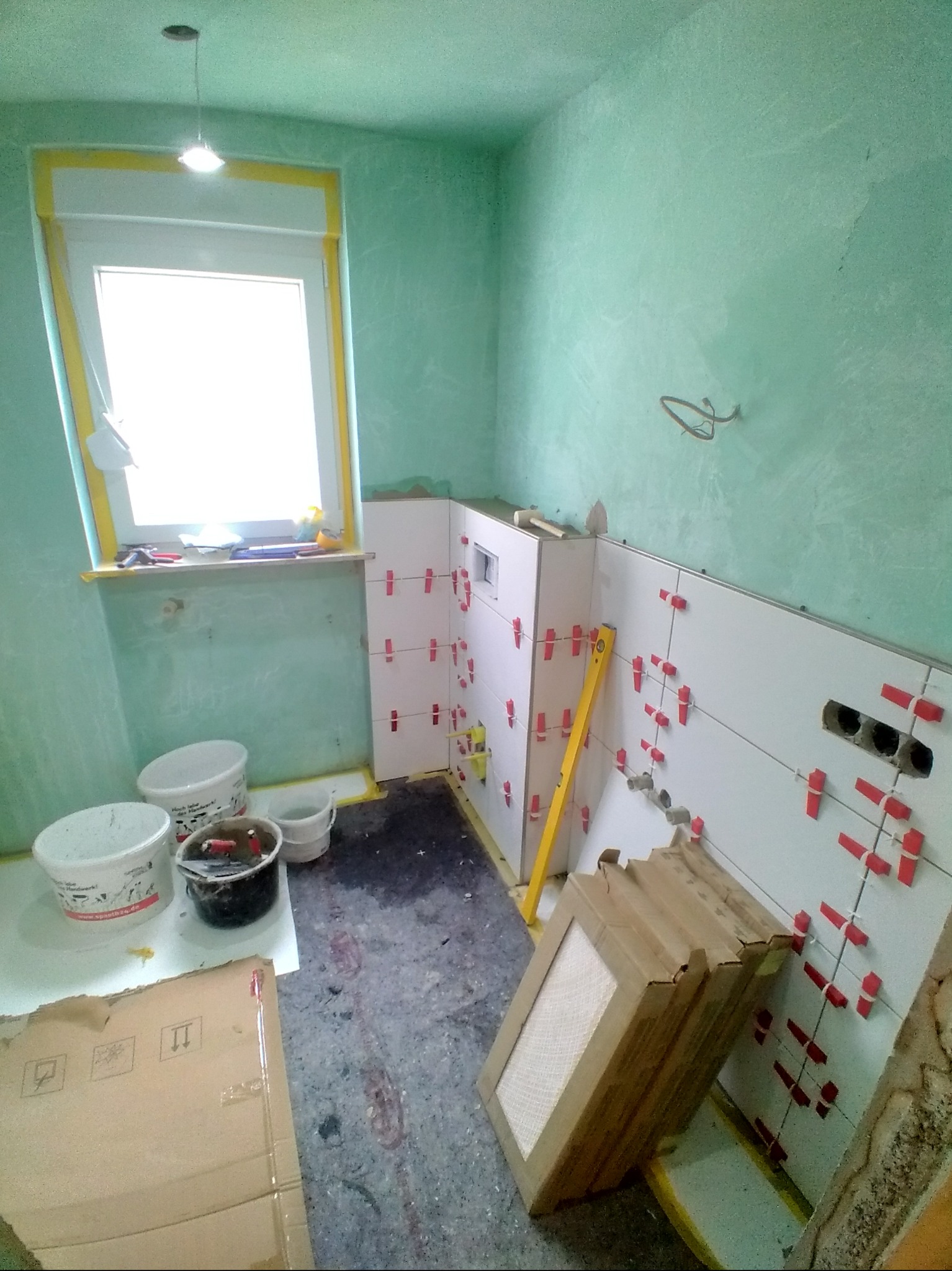 Łazienka w trakcie układania białych płytek z użyciem czerwonych krzyżyków dystansowych. Widoczna poziomica, puszki elektryczne i narzędzia glazurnicze.