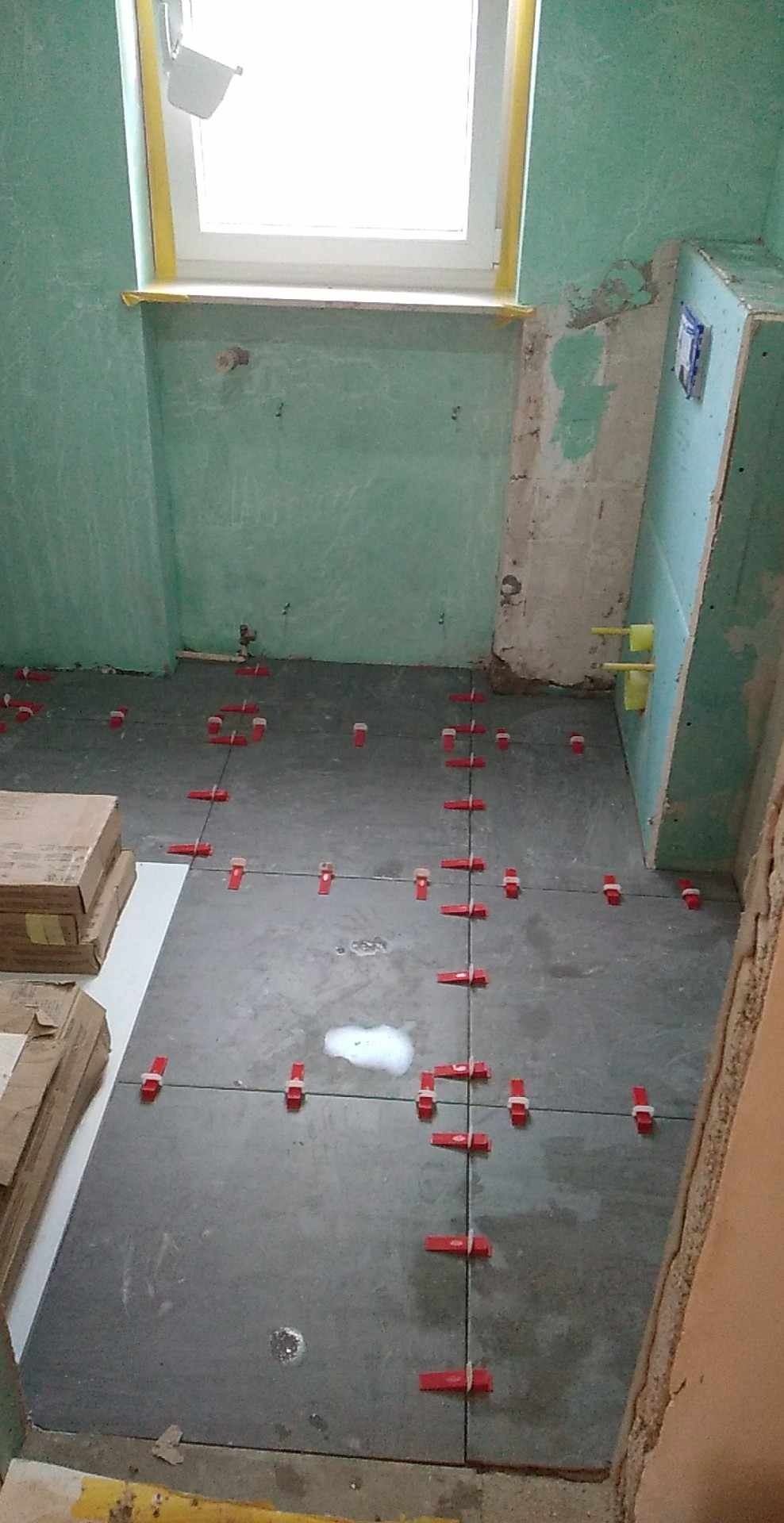 Układanie szarych płytek podłogowych w łazience z użyciem czerwonych klipsów poziomujących. Widoczne surowe ściany i okno z żółtą taśmą malarską.