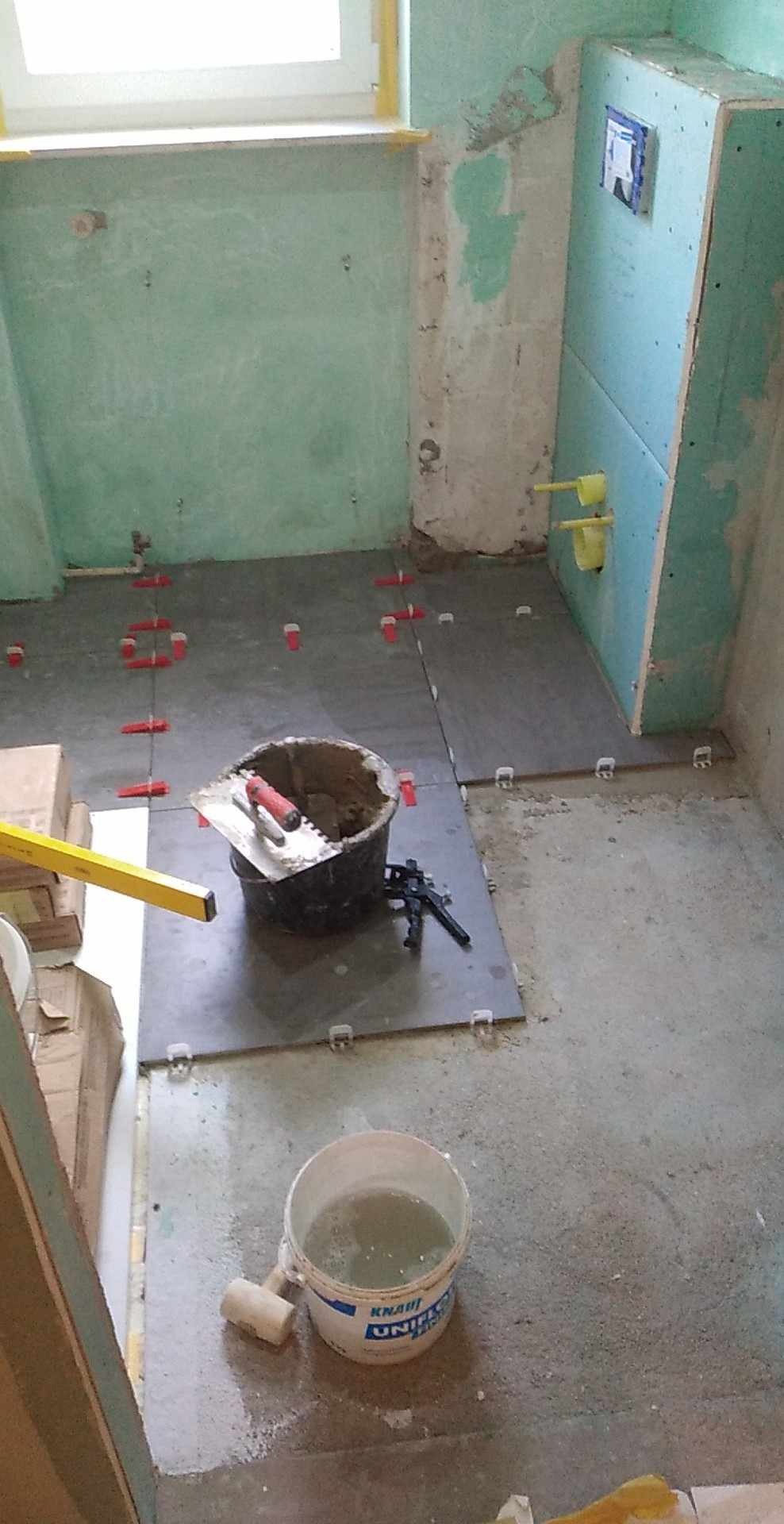 Układanie płytek podłogowych w łazience w trakcie remontu. Widoczne narzędzia, poziomica, wiadro z klejem i płytki z czerwonymi krzyżykami dystansowymi.