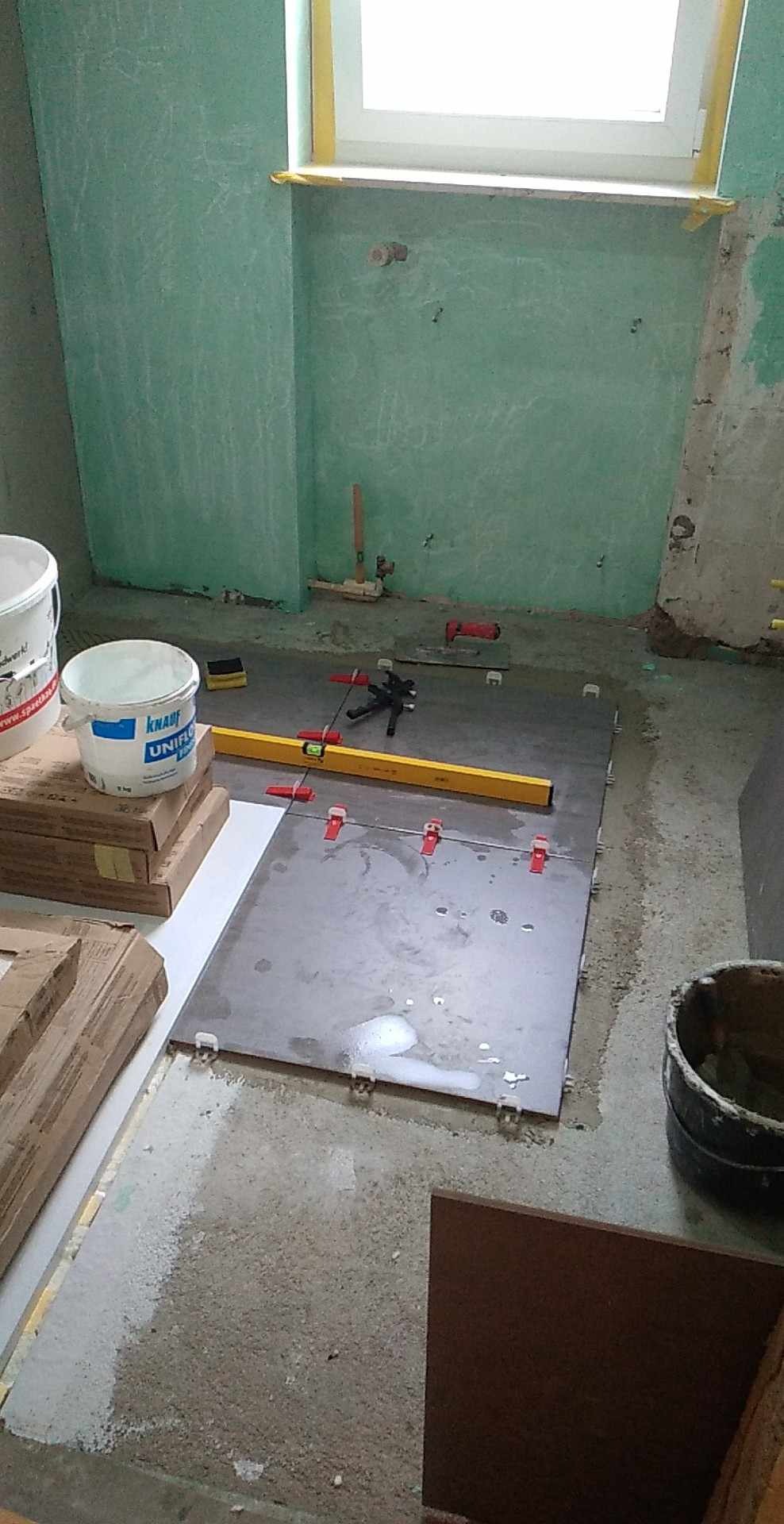 Układanie płytek podłogowych w łazience w trakcie remontu. Widoczne poziomica, krzyżyki dystansowe i wiadro z zaprawą. Ściany częściowo przygotowane pod glazurę.