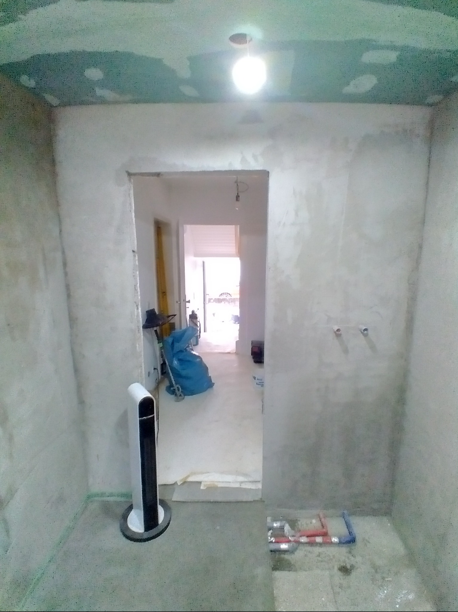 Surowa łazienka w trakcie prac: ściany po gładzi, widoczne rury instalacji wodnej, wentylator i narzędzia. Sufit z widocznymi śladami szpachlowania. Oświetlenie sufitowe.