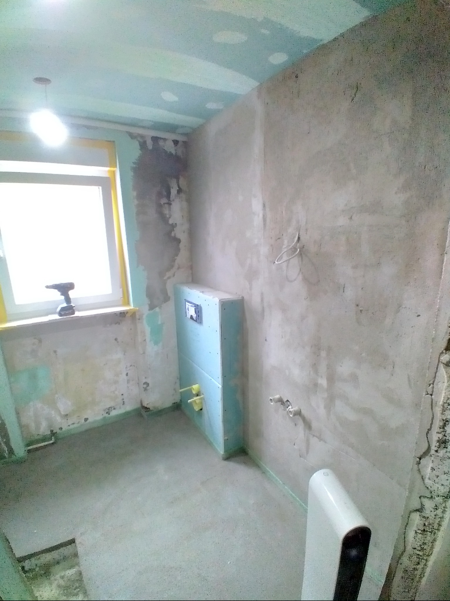 Łazienka w trakcie remontu: surowe ściany, zabudowa stelaża WC, okno z wkrętarką. Widoczne ślady po instalacjach i przygotowania do dalszych prac wykończeniowych.