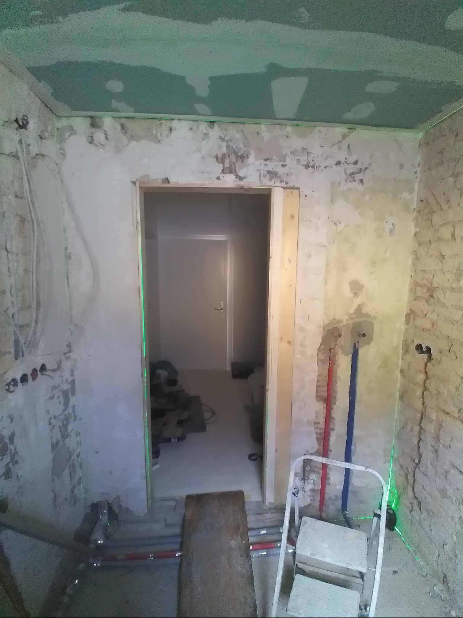 Remont łazienki w trakcie: surowe ściany, instalacja wodna (czerwona i niebieska rura), laser krzyżowy, przygotowany otwór na drzwi. Sufit z płyt karton-gips.