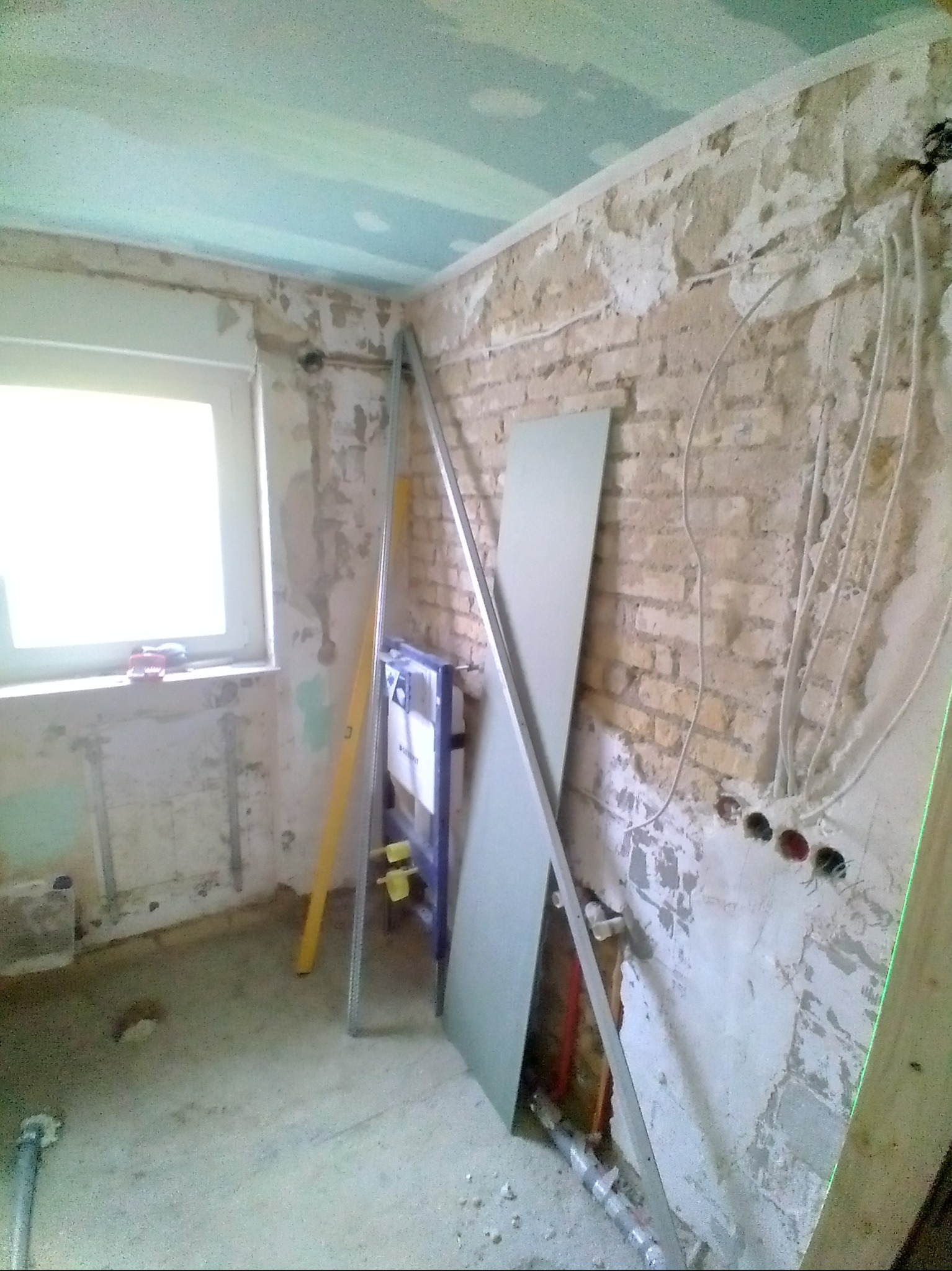 Wnętrze łazienki w trakcie remontu: odsłonięta cegła, instalacja podtynkowa, płyta gipsowa oparta o ścianę, stelaż WC. Widoczne przewody elektryczne i poziomica.