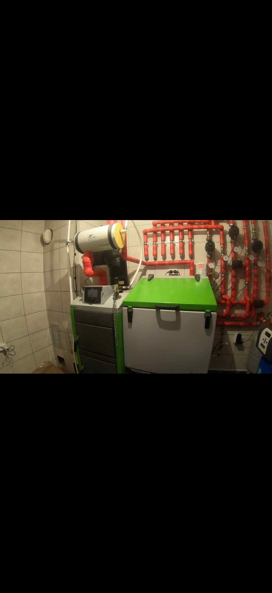 Kotłownia z piecem na pellet i zasobnikiem, widoczne czerwone rury instalacji grzewczej na ścianie z jasnymi płytkami. Ujęcie szerokokątne.