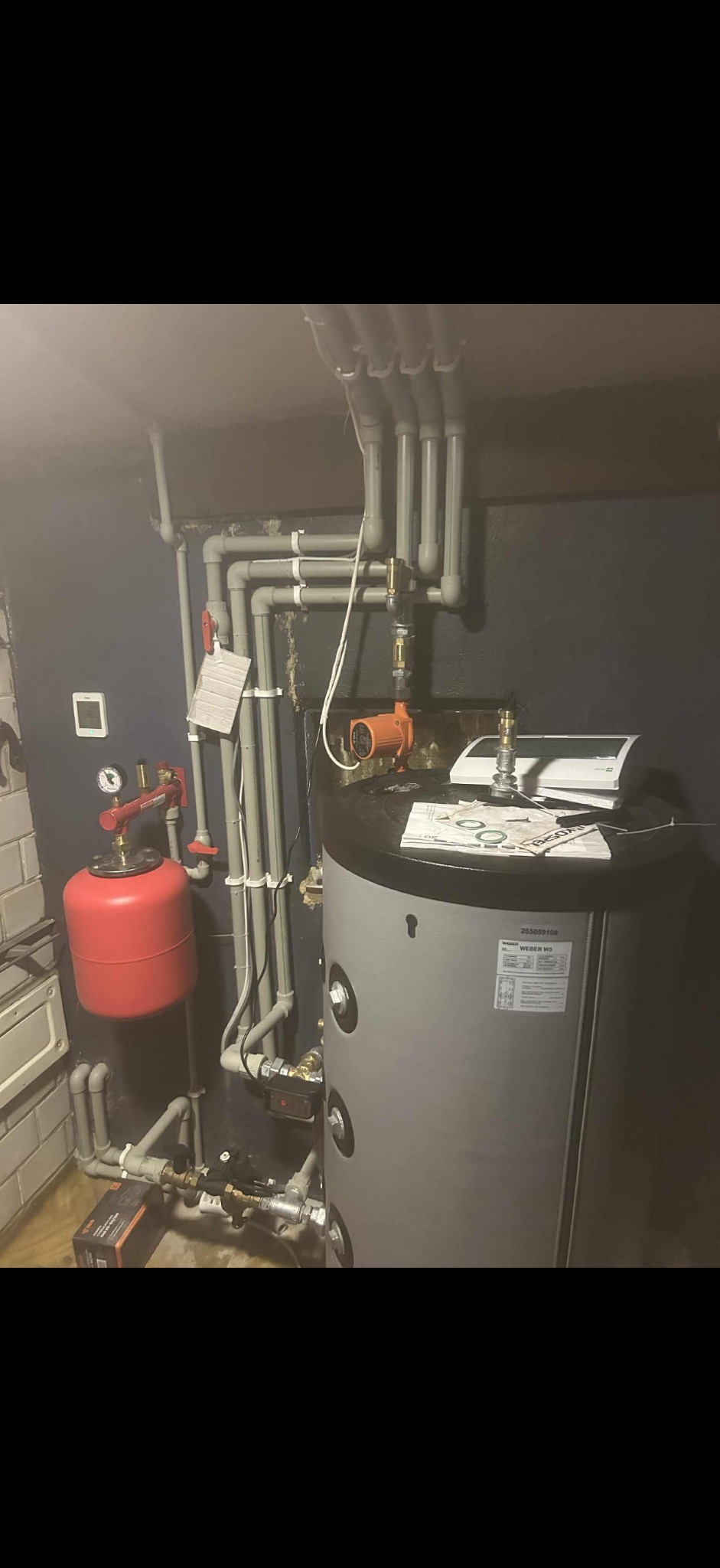 Szary bojler z podłączoną instalacją hydrauliczną i pompą obiegową w kotłowni. Widoczne rury, zawory, manometr i zbiornik wyrównawczy.