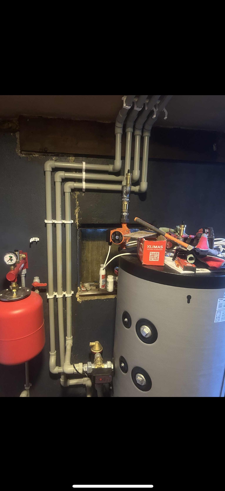 Szare rury instalacji CO/CWU z czerwonym zbiornikiem wyrównawczym i szarym zasobnikiem. Widoczne narzędzia i pudełko Klimas. Instalacja w ciemnym pomieszczeniu.