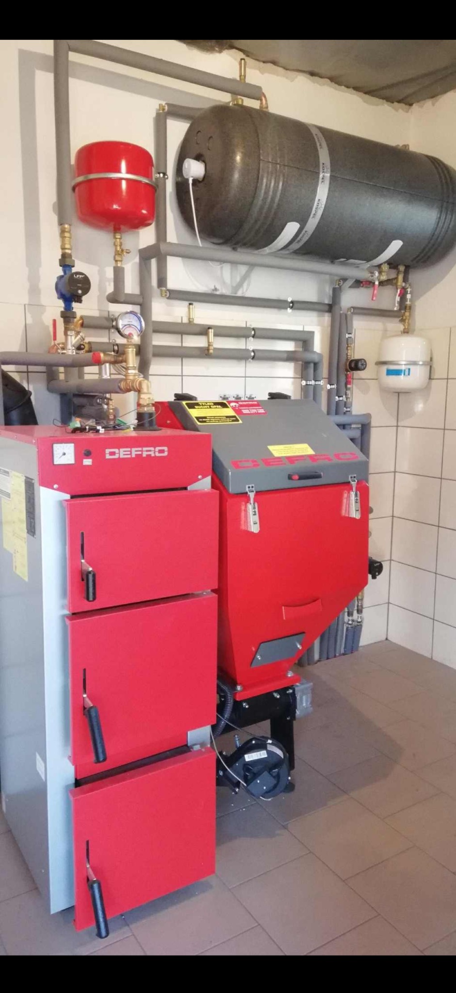 Czerwono-szary piec Defro z instalacją grzewczą w kotłowni z białymi płytkami. Widoczne rury, zbiorniki wyrównawcze i manometry. Oświetlenie górne.