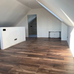 Evibud - Jasne poddasze po remoncie z panelami podłogowymi w odcieniach brązu. Widoczne białe ściany, okno dachowe i fragment ściany działowej z gniazdkami.