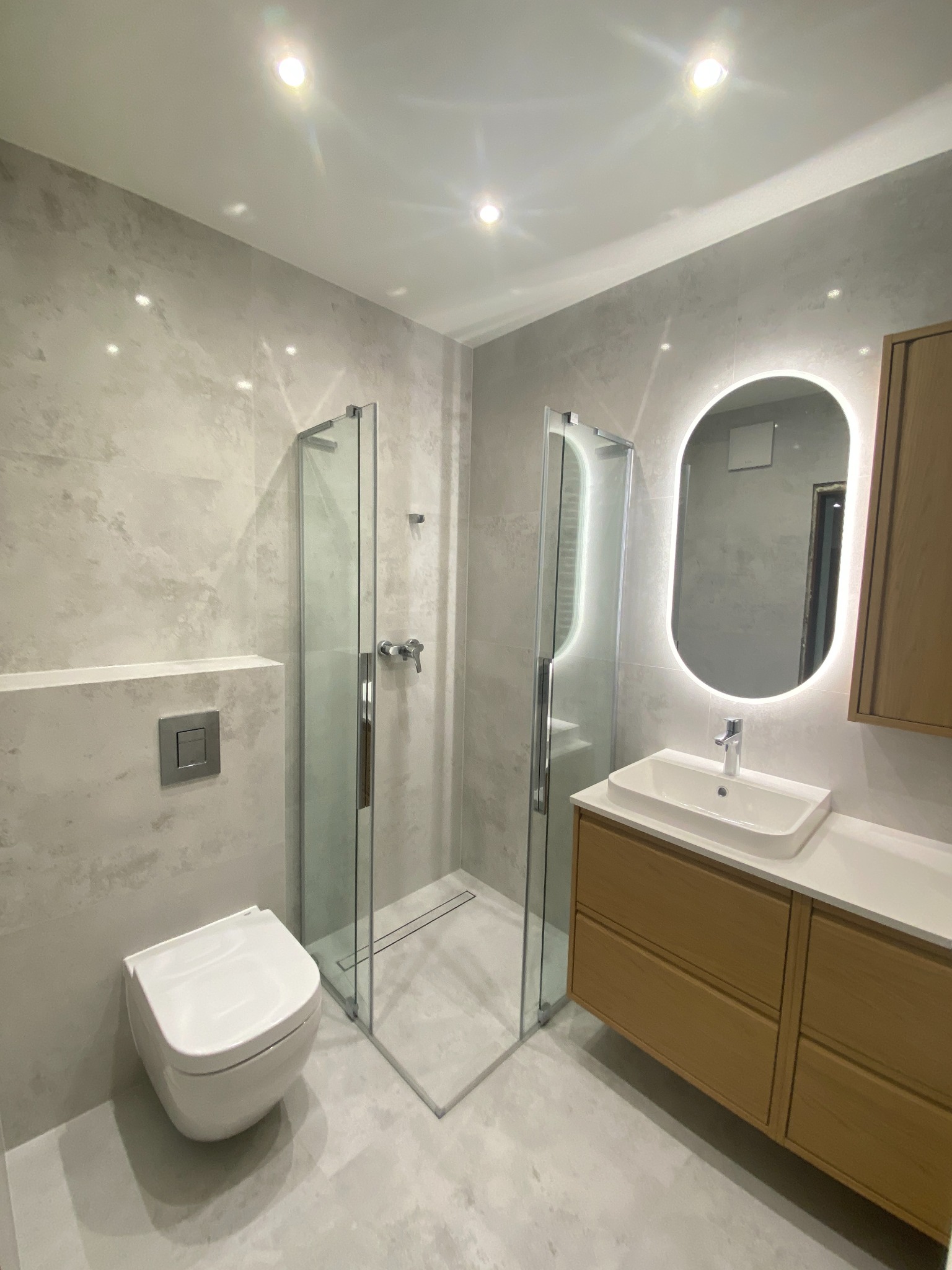 Nowoczesna łazienka z kabiną prysznicową typu walk-in, podwieszaną toaletą i szafką z umywalką podświetlaną owalnym lustrem. Szare płytki imitujące beton.