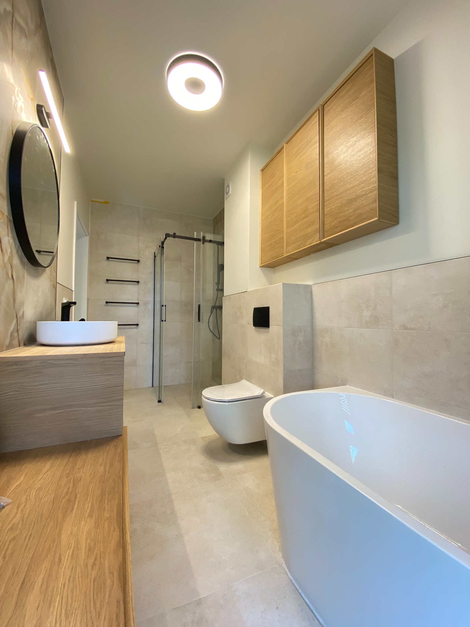 Nowoczesna łazienka z drewnianymi akcentami: szafka nad toaletą, umywalka na blacie, okrągłe lustro i wolnostojąca wanna. Beżowe płytki i kabina prysznicowa.