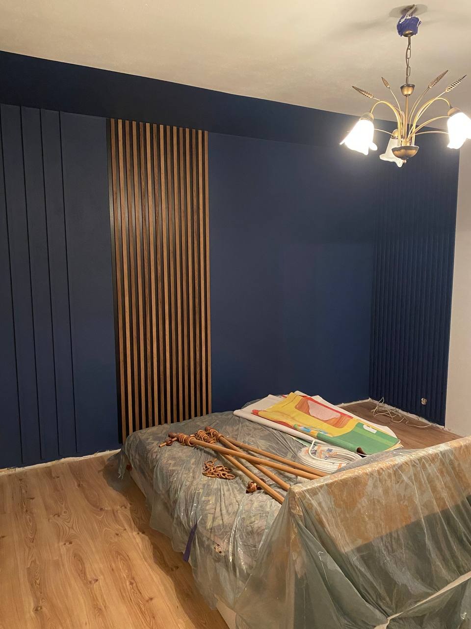 Granatowa ściana z drewnianymi lamelami i listwami, złoty żyrandol. Częściowo zakryte łóżko folią malarską. Widoczne drewniane elementy dekoracyjne.