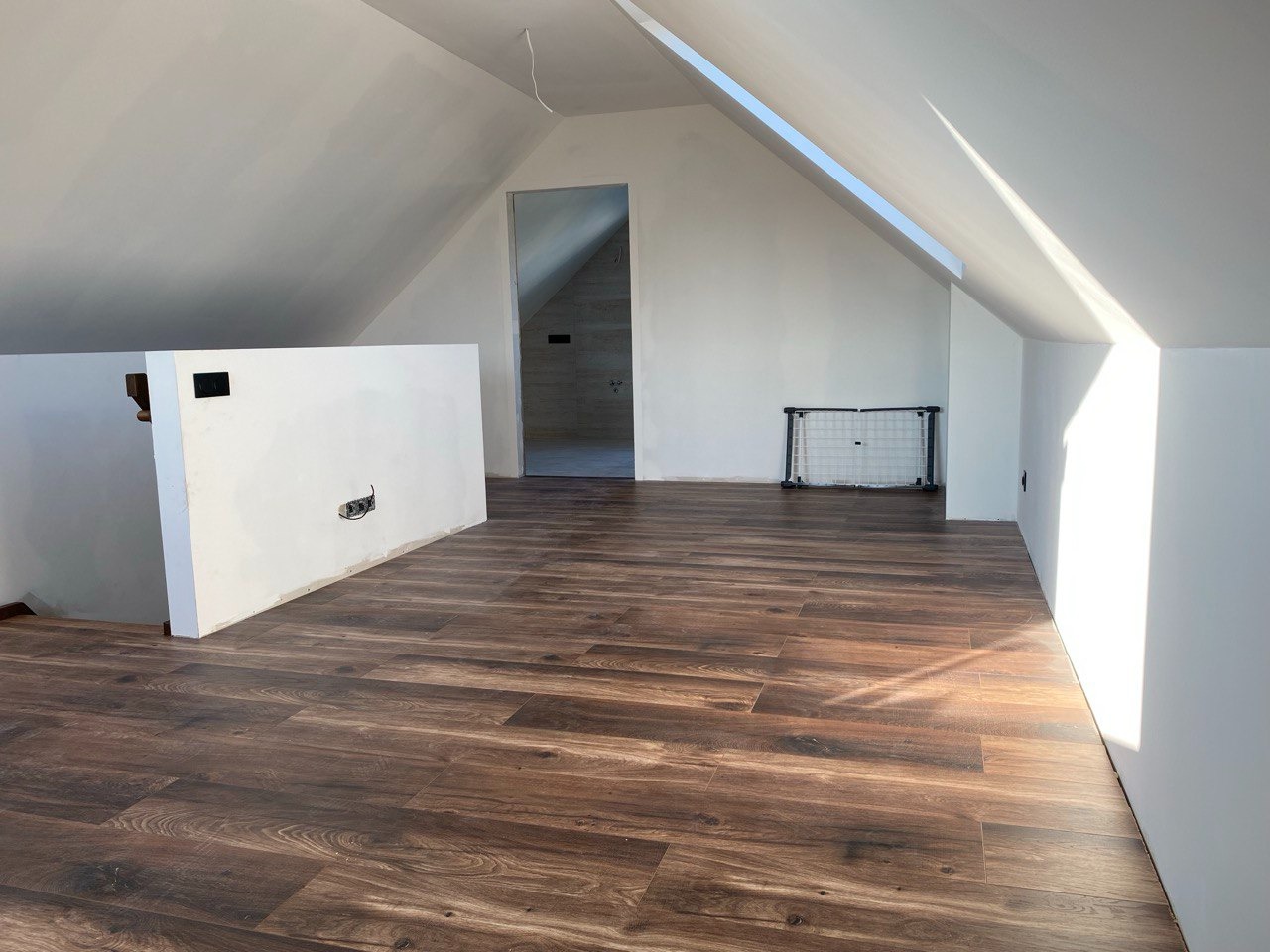 Jasne poddasze po remoncie z panelami podłogowymi w odcieniach brązu. Widoczne białe ściany, okno dachowe i fragment ściany działowej z gniazdkami.