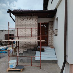 POLBUD PAWEŁ GLISZCZYŃSKI - Wykończenie elewacji wejściowej domu z użyciem cegieł klinkierowych, widoczne rusztowanie i materiały budowlane. Częściowo wykończone schody wejściowe.