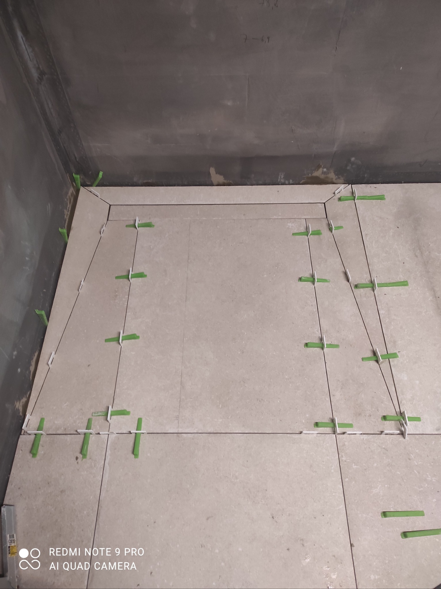 Układanie jasnych płytek podłogowych w narożnym pomieszczeniu, z użyciem zielonych krzyżyków dystansowych. Widoczne fugi i surowa ściana z notatkami.