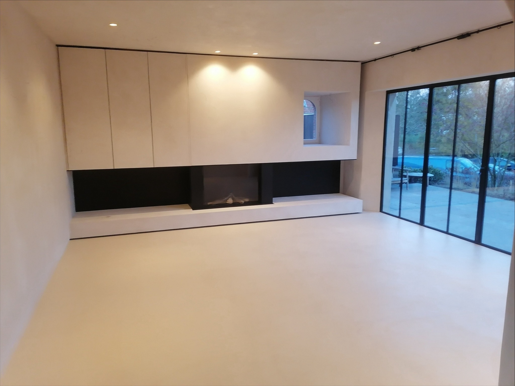 Minimalistyczne wnętrze: gładkie, beżowe ściany i podłoga, zabudowa meblowa z czarnymi wnękami, duże okno z czarną ramą. Nowoczesny design.