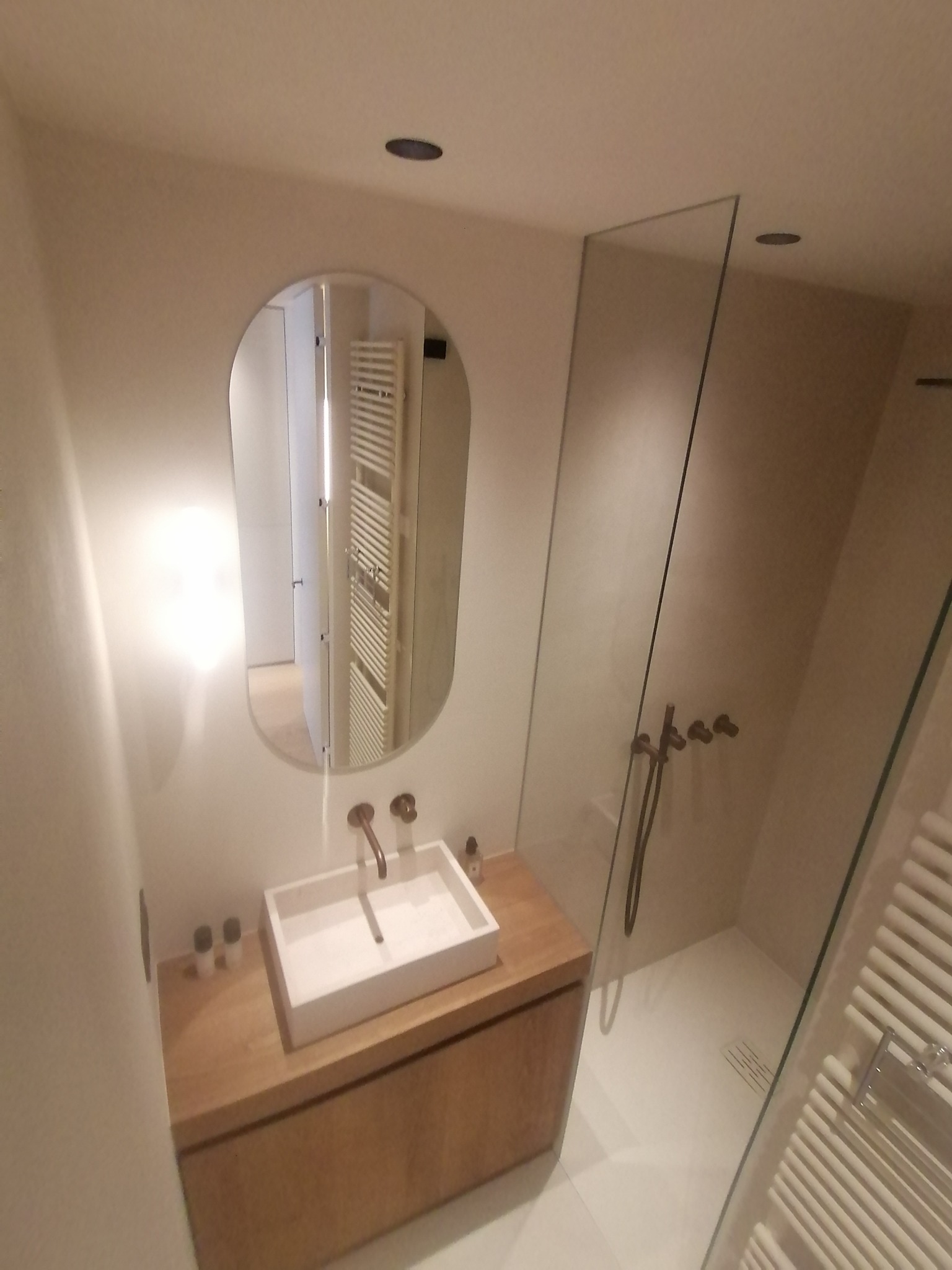 Nowoczesna łazienka z owalnym lustrem, białą umywalką na drewnianej szafce i kabiną prysznicową ze szklaną ścianką. Minimalistyczny design, ciepłe oświetlenie.