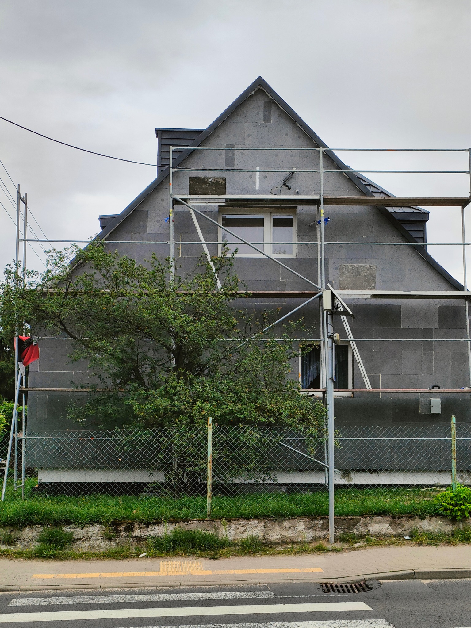 Dom w trakcie ocieplania, elewacja pokryta szarym styropianem, rusztowania, widok z ulicy, pochmurne niebo. Prace wykończeniowe na zewnątrz budynku.