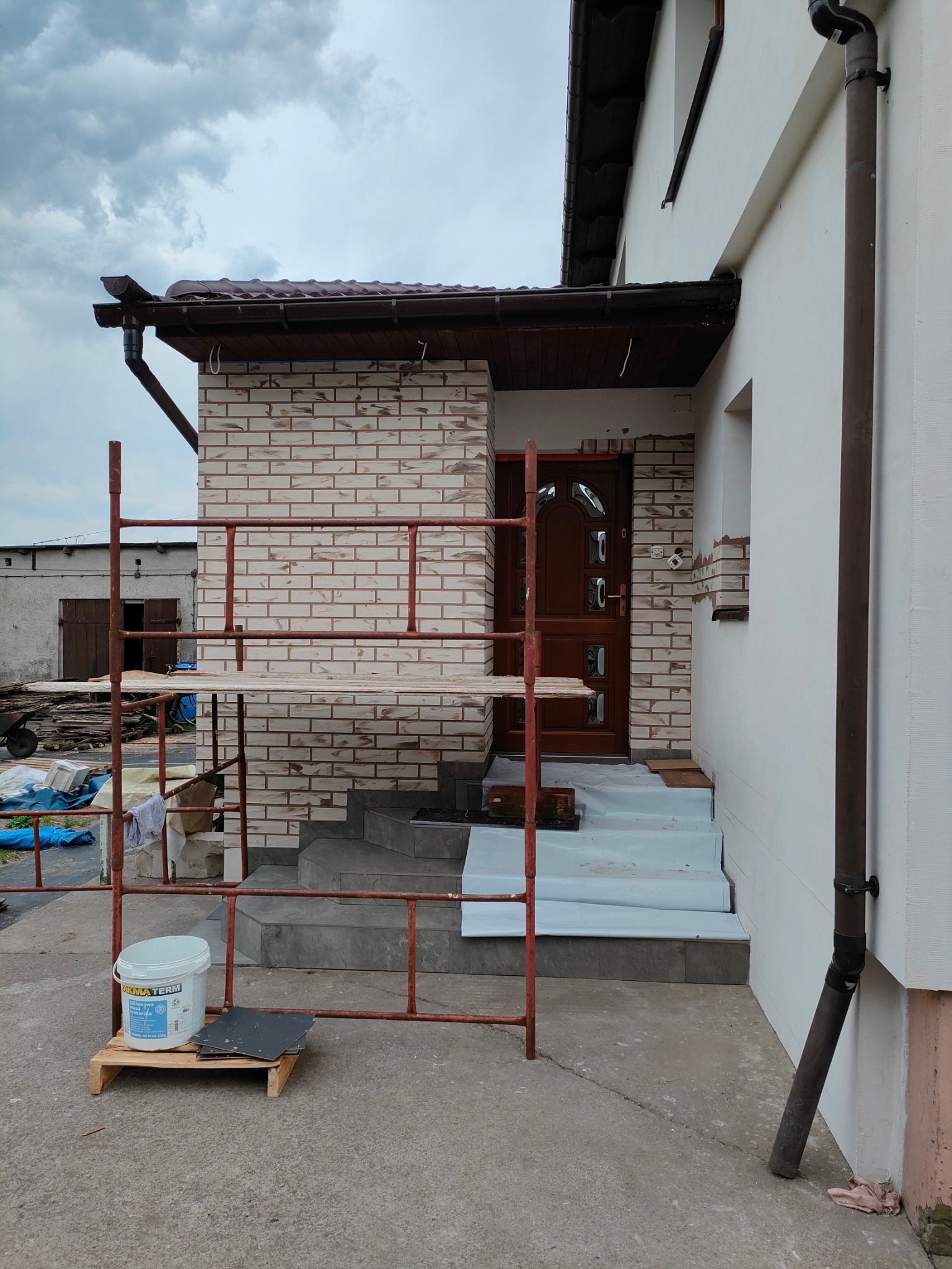 Wykończenie elewacji wejściowej domu z użyciem cegieł klinkierowych, widoczne rusztowanie i materiały budowlane. Częściowo wykończone schody wejściowe.