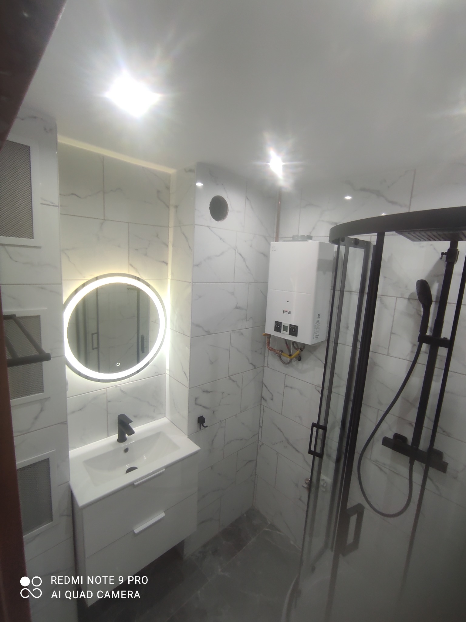 Nowoczesna łazienka z marmurowymi płytkami, okrągłym lustrem z podświetleniem LED, białą szafką pod umywalką i czarną kabiną prysznicową.