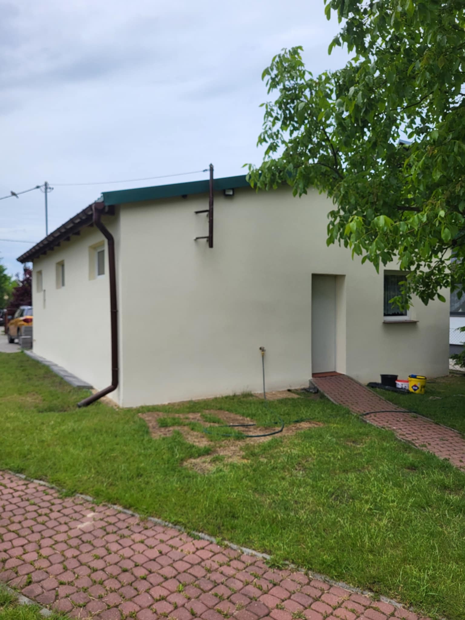 Otynkowany budynek gospodarczy z brązowymi rynnami i chodnikiem z kostki, widok z zewnątrz w pochmurny dzień. Widoczne wiadro z farbą.