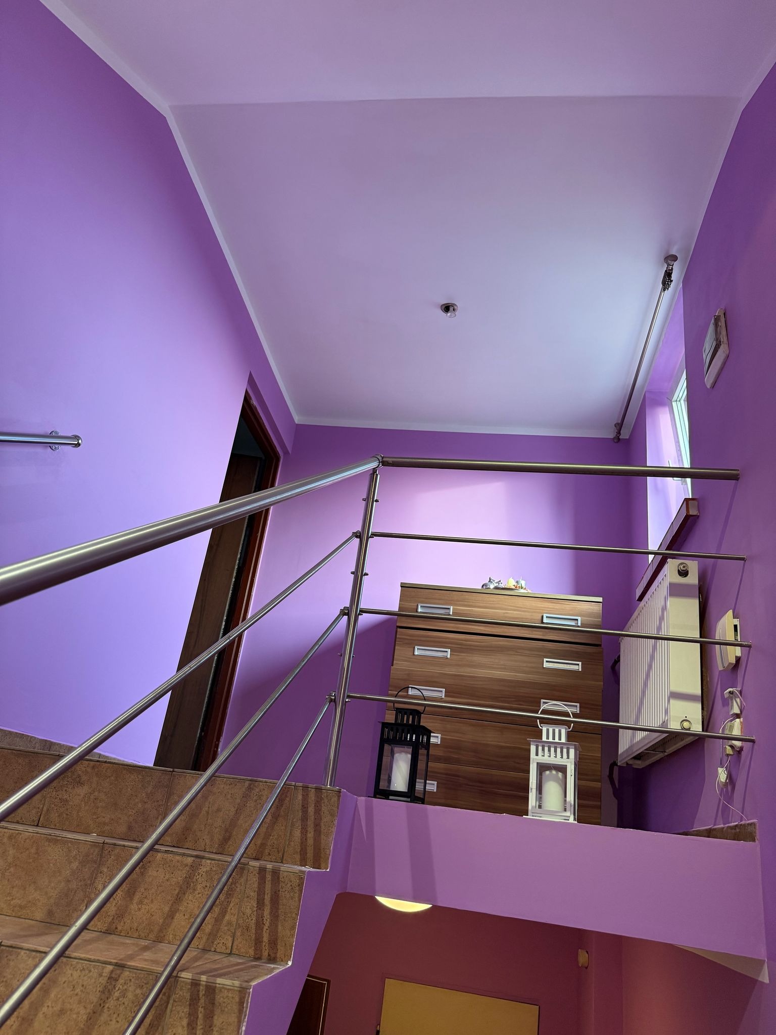 Fioletowe ściany klatki schodowej z brązowymi schodami i metalową barierką. Na podeście komoda i lampiony. Widok z dołu, ukośny kadr.
