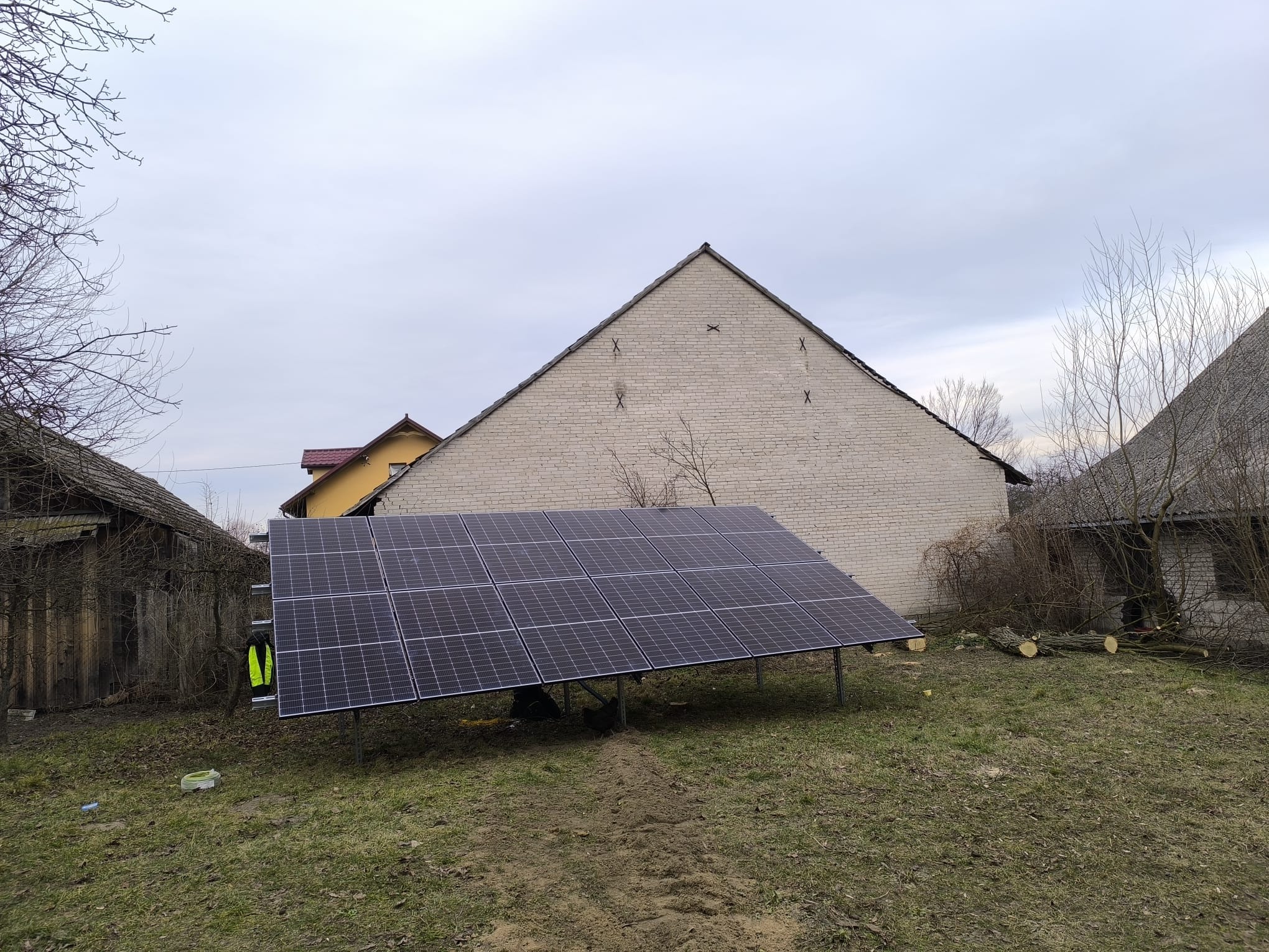 Panel fotowoltaiczny na metalowej konstrukcji, ustawiony na trawiastym terenie przed budynkiem z cegły. Widoczna kamizelka odblaskowa.