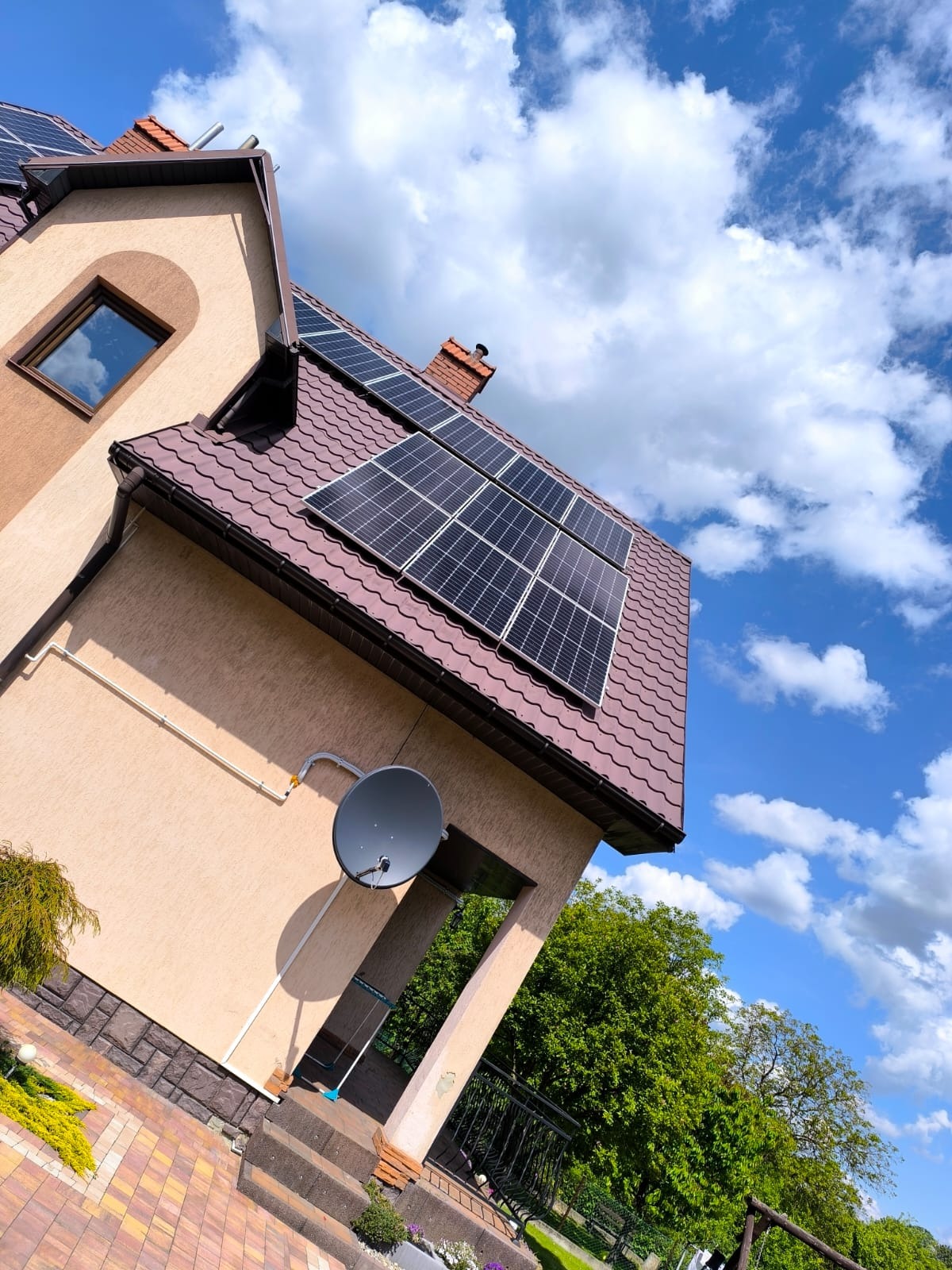 Dom jednorodzinny z brązowym dachem i panelami fotowoltaicznymi na tle błękitnego nieba z chmurami. Na ścianie budynku widoczna antena satelitarna.