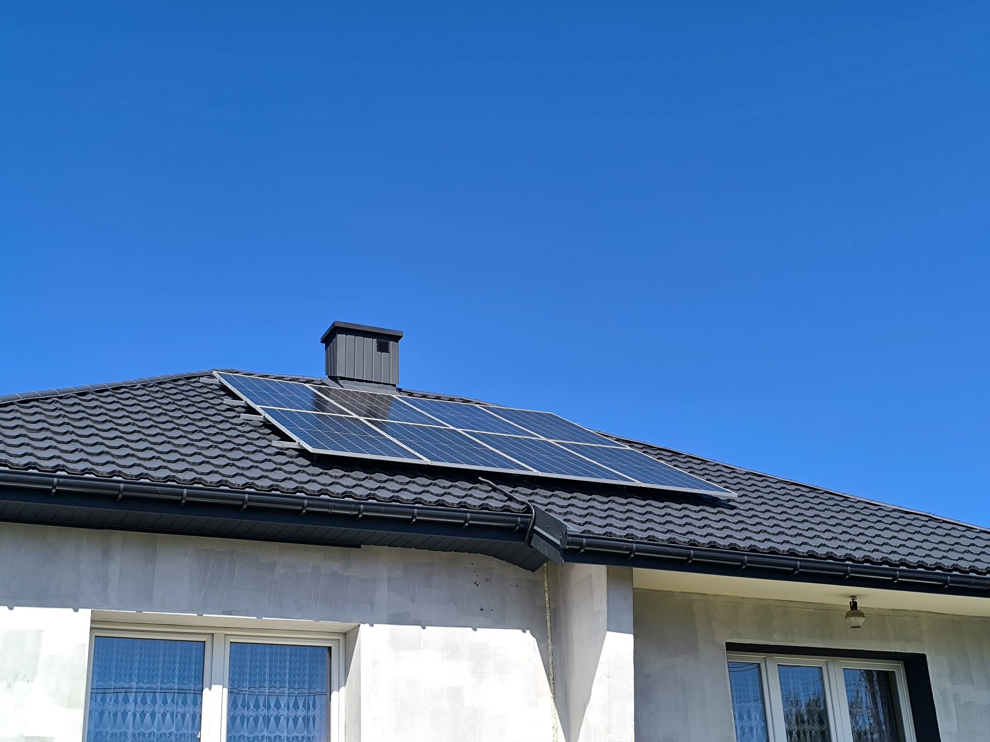 Panele fotowoltaiczne na dachu domu z szarej dachówki, widoczne okna z białymi ramami i błękitne niebo w tle. Dom w stanie surowym.