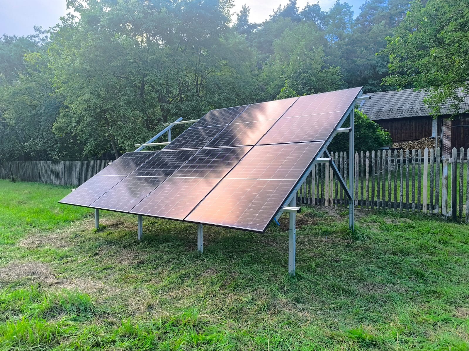 Panel fotowoltaiczny na metalowej konstrukcji, ustawiony na trawie przed drewnianym płotem i budynkiem w tle, w słoneczny dzień. Widok z boku.
