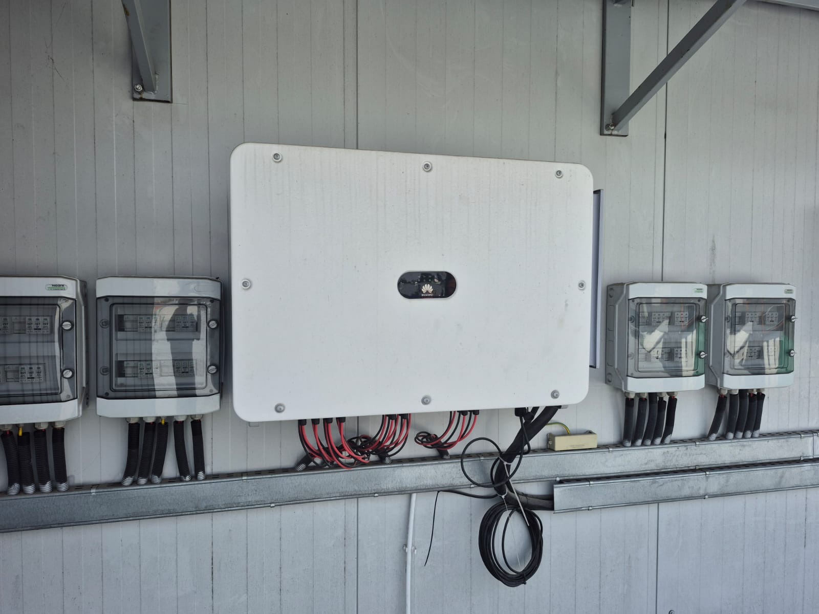 Biały falownik Huawei z podłączonymi przewodami i zabezpieczeniami na metalowej szynie, zamontowany na ścianie z paneli. Widoczne skrzynki z bezpiecznikami.