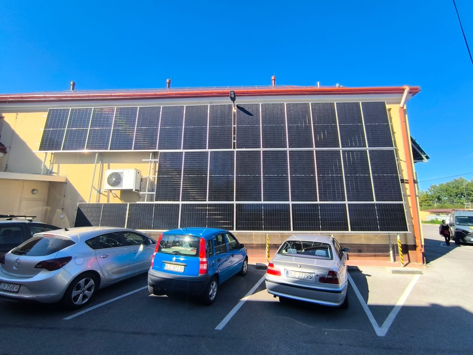 Elewacja budynku z panelami fotowoltaicznymi zamontowanymi na całej ścianie, widoczne zaparkowane samochody i błękitne niebo.