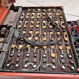 DB ElektroSystem - Otwarta komora baterii wózka widłowego z widocznymi licznymi ogniwami i przewodami. W tle narzędzia i fragment samochodu.