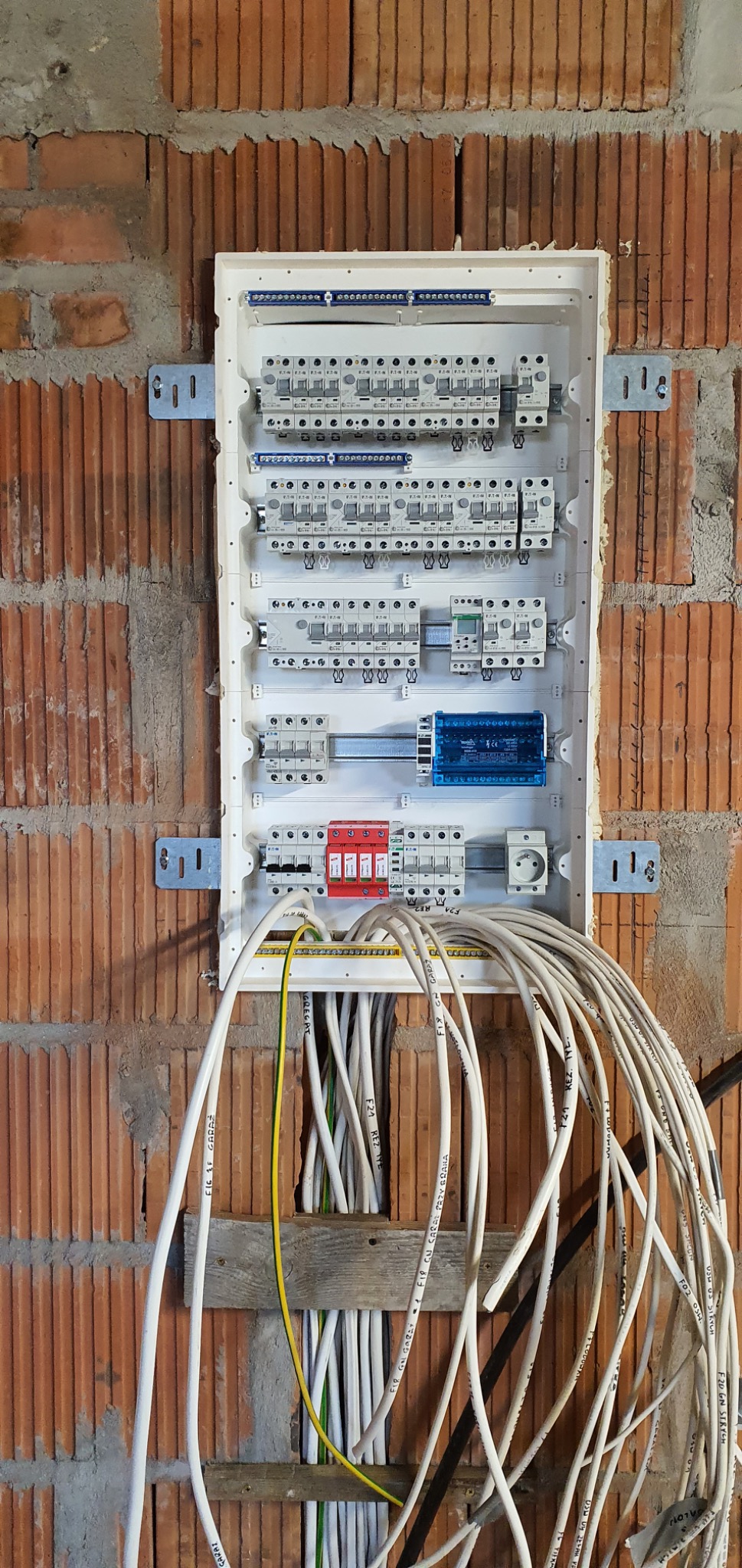 Rozdzielnia elektryczna z zabezpieczeniami i przewodami, zamontowana na ceglanej ścianie. Widoczne bezpieczniki, wyłączniki różnicowoprądowe i przewody elektryczne.