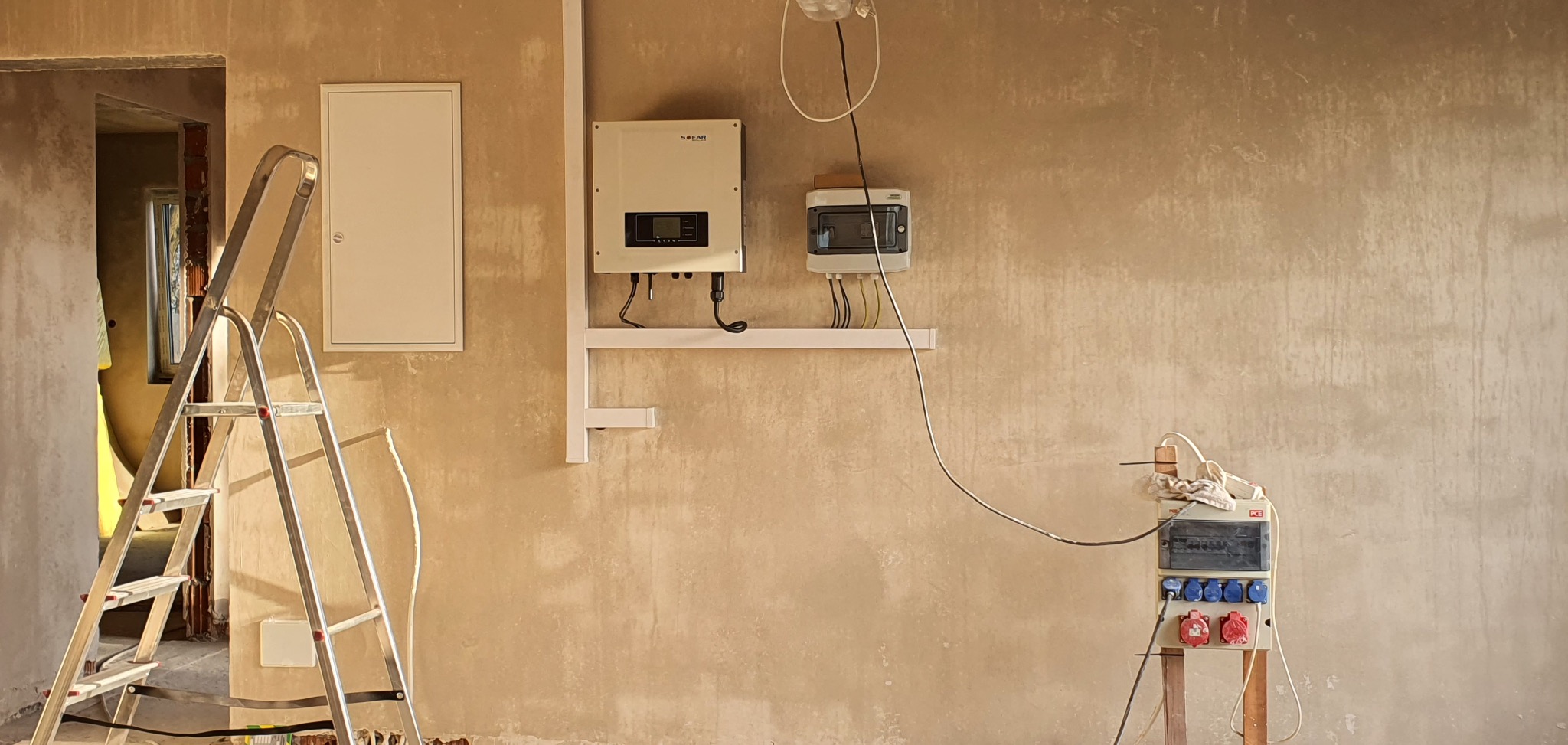 Wnętrze w trakcie prac: rozdzielnica elektryczna, inwerter solarny i skrzynka bezpiecznikowa zamontowane na ścianie. Obok drabina aluminiowa.