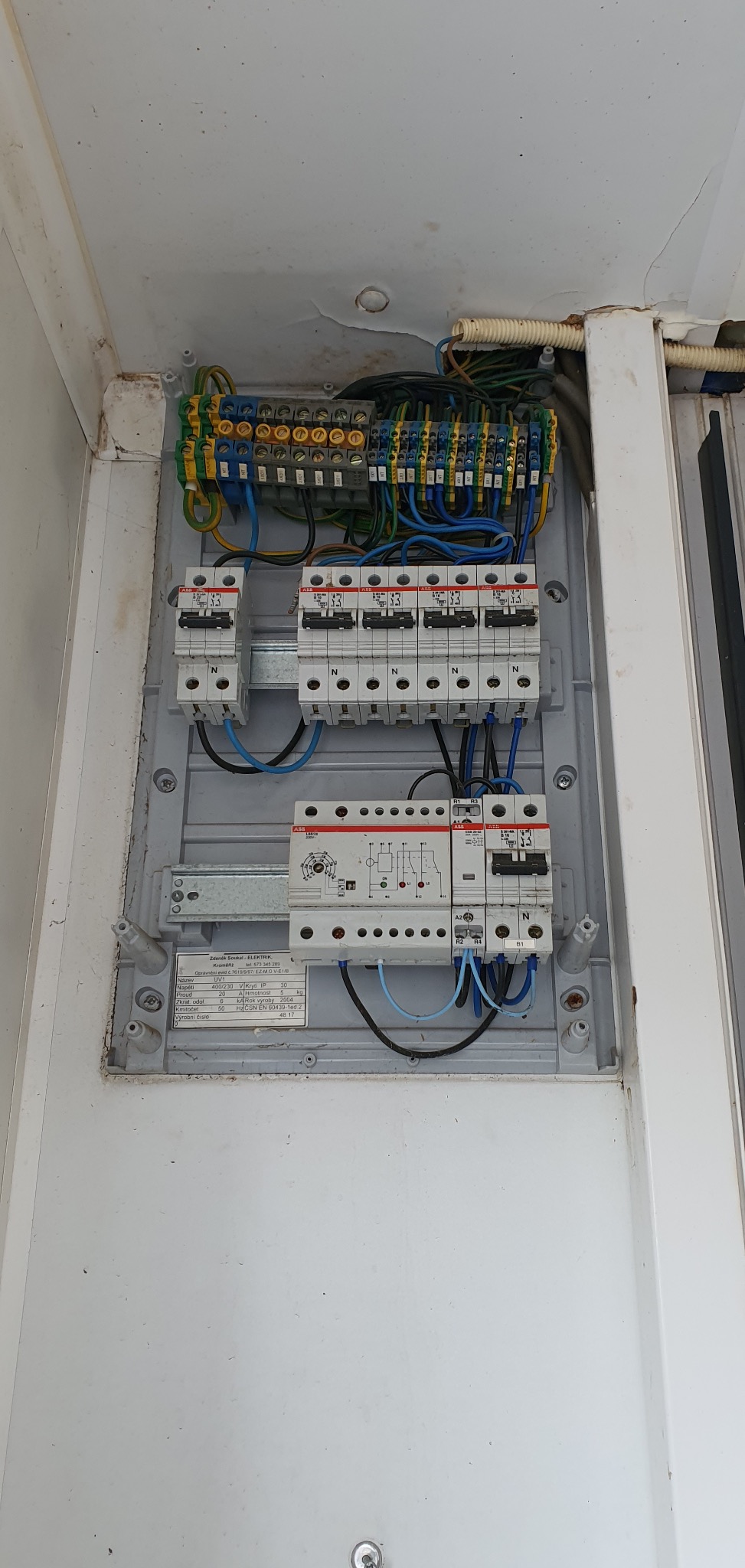 Szara skrzynka elektryczna z bezpiecznikami i przewodami wewnątrz, widoczne złącza i oznaczenia. Instalacja wewnątrz obudowy.