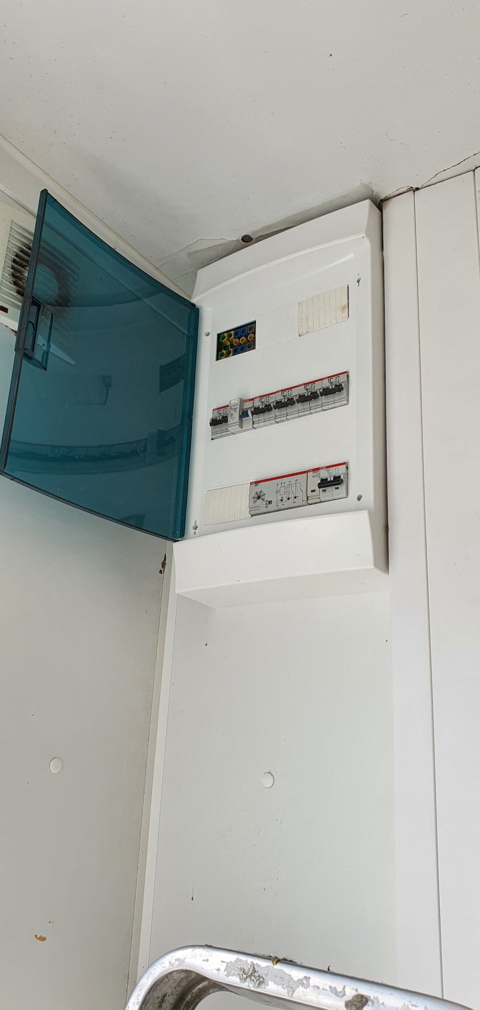 Otwarta skrzynka elektryczna z bezpiecznikami na białej ścianie, widoczne okablowanie i elementy instalacji. Niebieska osłona skrzynki uchylona.