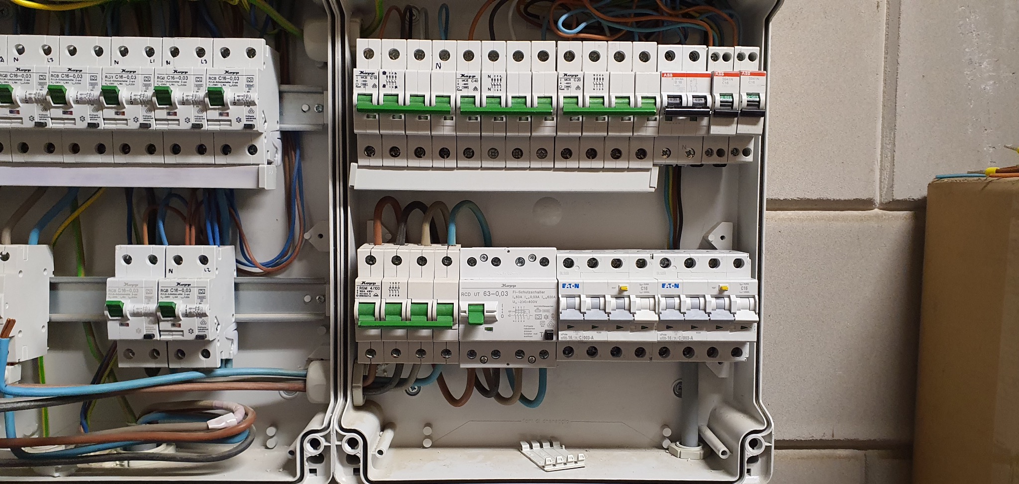 Rozdzielnica elektryczna z zainstalowanymi bezpiecznikami różnych marek (Kopp, ABB, Eaton) i okablowaniem, widok z bliska na ścianie z cegieł.