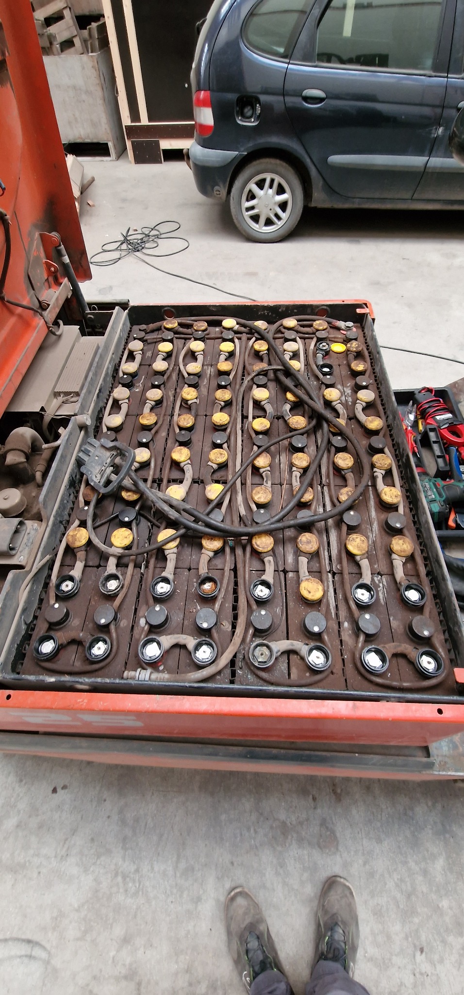 Otwarta komora baterii wózka widłowego z widocznymi licznymi ogniwami i przewodami. W tle narzędzia i fragment samochodu.