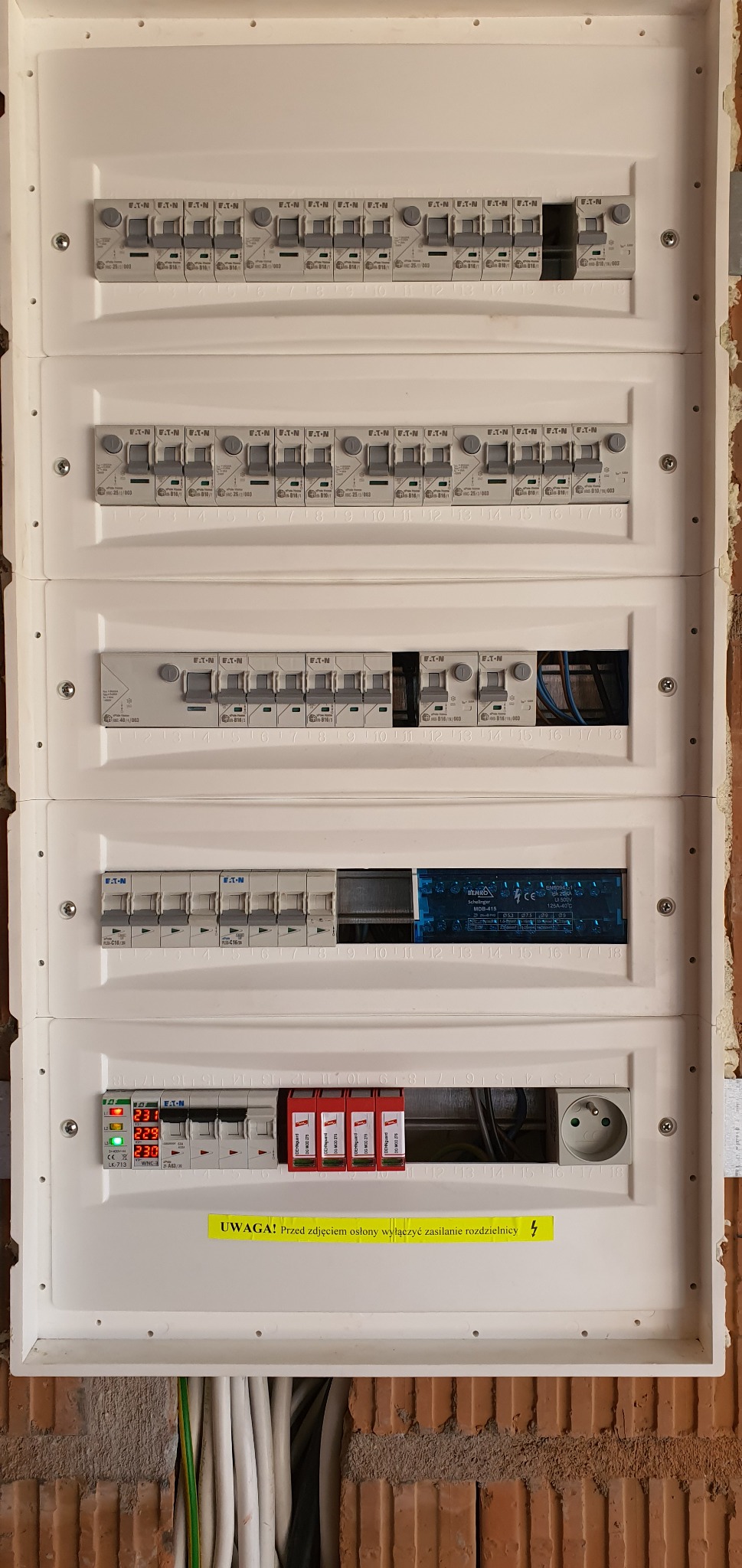 Rozdzielnica elektryczna Eaton z zabezpieczeniami, widoczne wyłączniki nadprądowe, ograniczniki przepięć i gniazdo. Żółta naklejka ostrzegawcza. Instalacja w trakcie montażu.