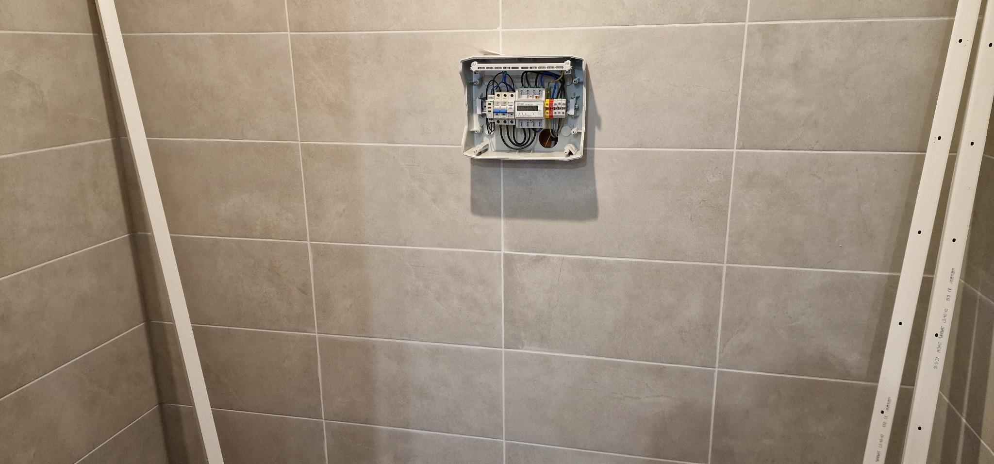 Otwarta skrzynka elektryczna z okablowaniem na kafelkowanej ścianie. Widoczne elementy instalacji i przewody. Po bokach listwy przypodłogowe.