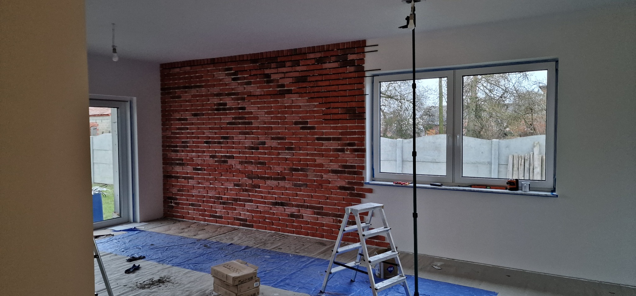 Wnętrze w trakcie wykańczania: ściana z cegły klinkierowej, drabina, poziomica przy oknie. Podłoga zabezpieczona folią malarską. Widoczne narzędzia i materiały budowlane.