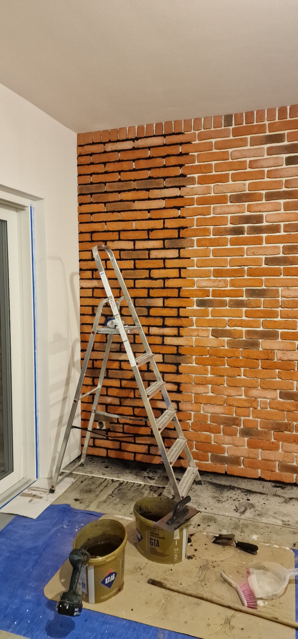 Ściana z cegły klinkierowej w trakcie fugowania. Część ściany już z czarną fugą, druga część jeszcze bez. Aluminiowa drabina oparta o ścianę. Wiadra z zaprawą i narzędzia.