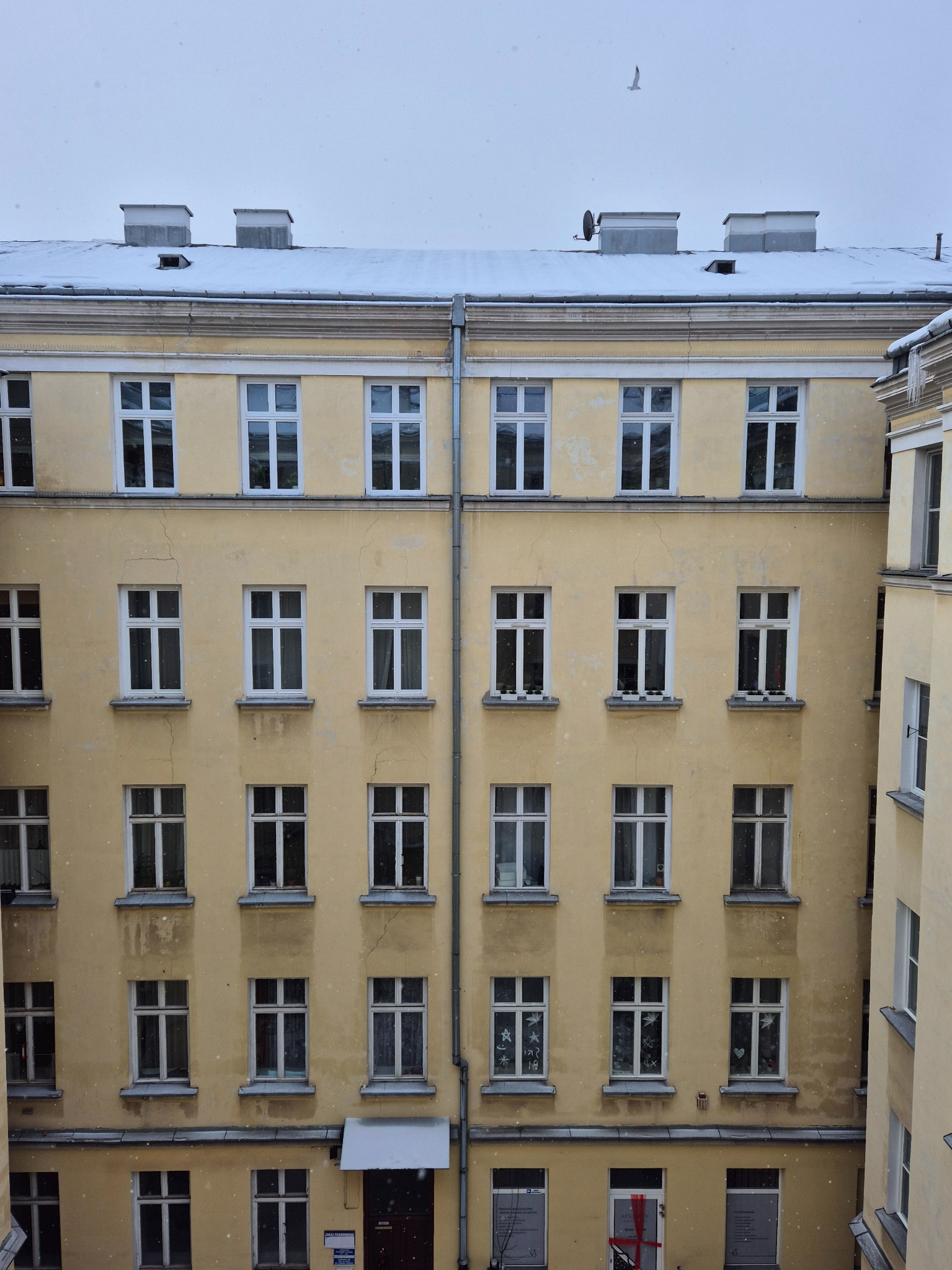 Żółta kamienica w zimowej scenerii, lekko ośnieżony dach, padający śnieg, widok z góry na budynek z licznymi oknami i elementami architektonicznymi.