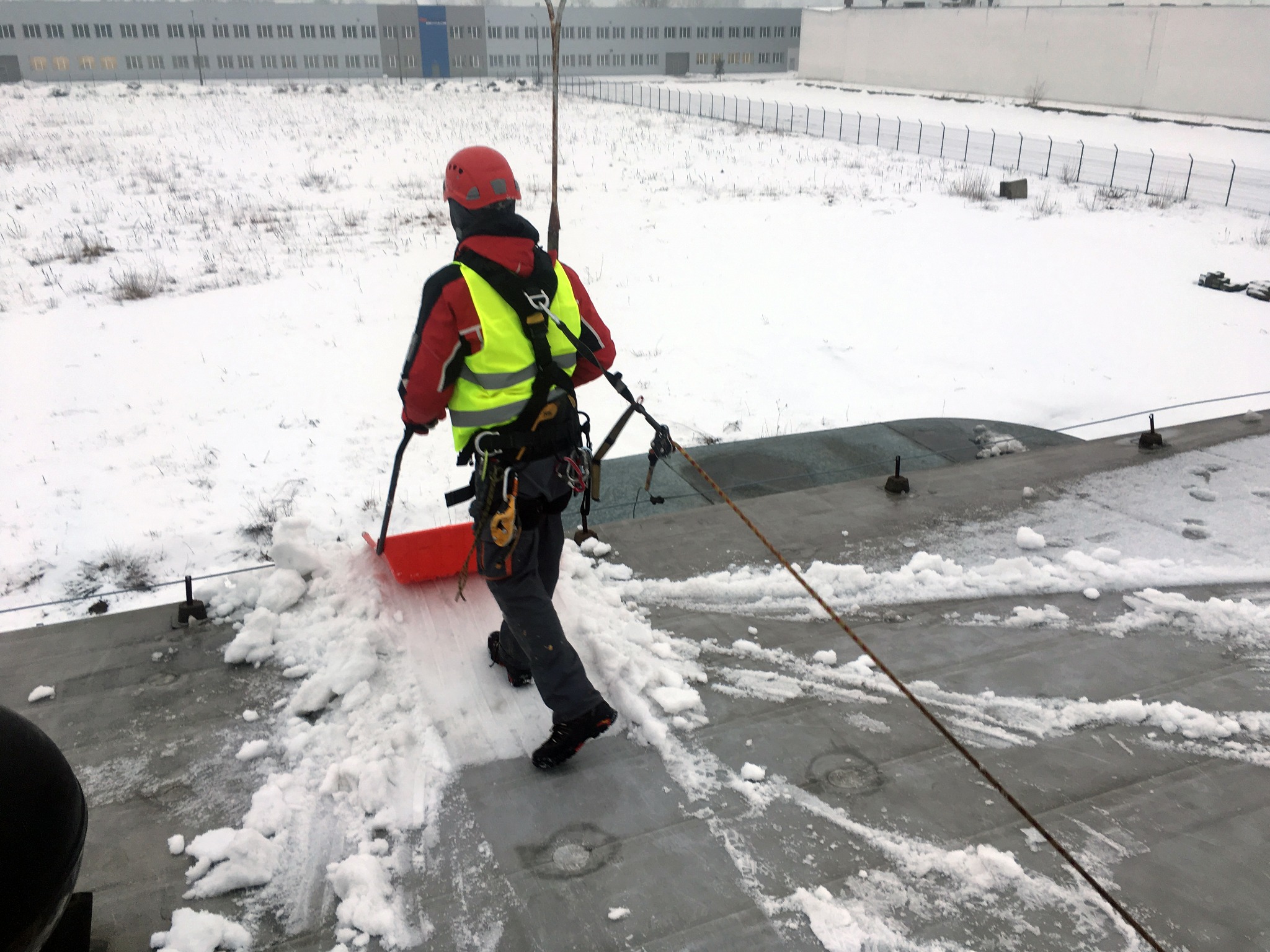 Pracownik w uprzęży, z czerwoną łopatą, odśnieża płaski dach budynku przemysłowego. W tle zaśnieżone pole i budynek.