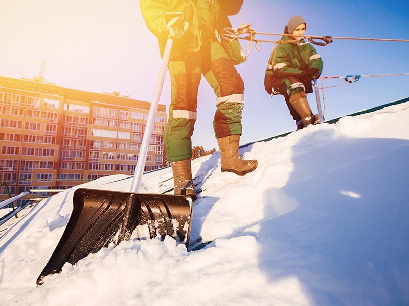 Dwóch pracowników w kombinezonach odśnieża dach budynku mieszkalnego za pomocą łopaty, zabezpieczonych linami. Słoneczny dzień, widok z dołu.