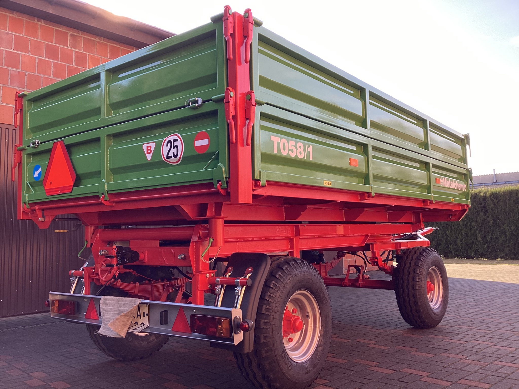 Zielono-czerwona przyczepa rolnicza T058/1 z tablicą rejestracyjną, widoczna z tyłu na tle ceglanego budynku i kostki brukowej.