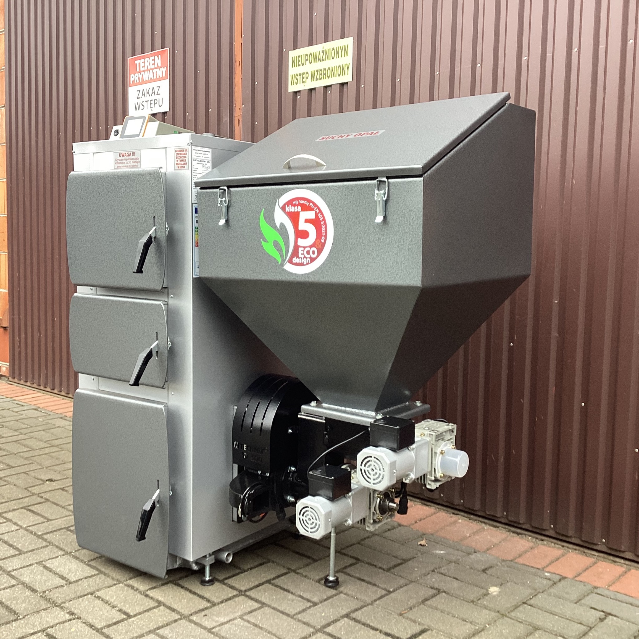 Szary piec na paliwo stałe klasy 5 ECO design, z widocznymi drzwiczkami i podajnikiem, ustawiony na zewnątrz budynku z brązową blachą trapezową w tle.